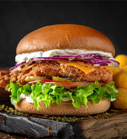 A crispy chicken sandwich with lettuce, sliced tomatoes, melted cheese, and red cabbage in a bun, on a rustic board with potatoes in the background.
