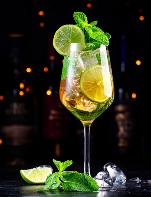 A cocktail in a wine glass garnished with lime slices and mint leaves sits on a dark table with ice cubes. Warm bokeh lights glow in the background.