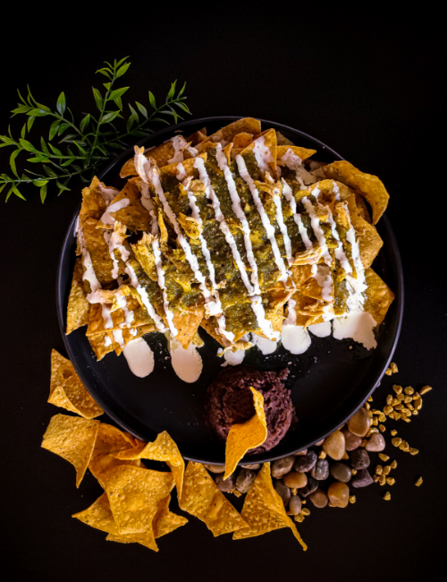 A plate of nachos topped with green sauce and white drizzles sits on a black surface. Garnished with a dollop of black beans and a side of corn kernels.
