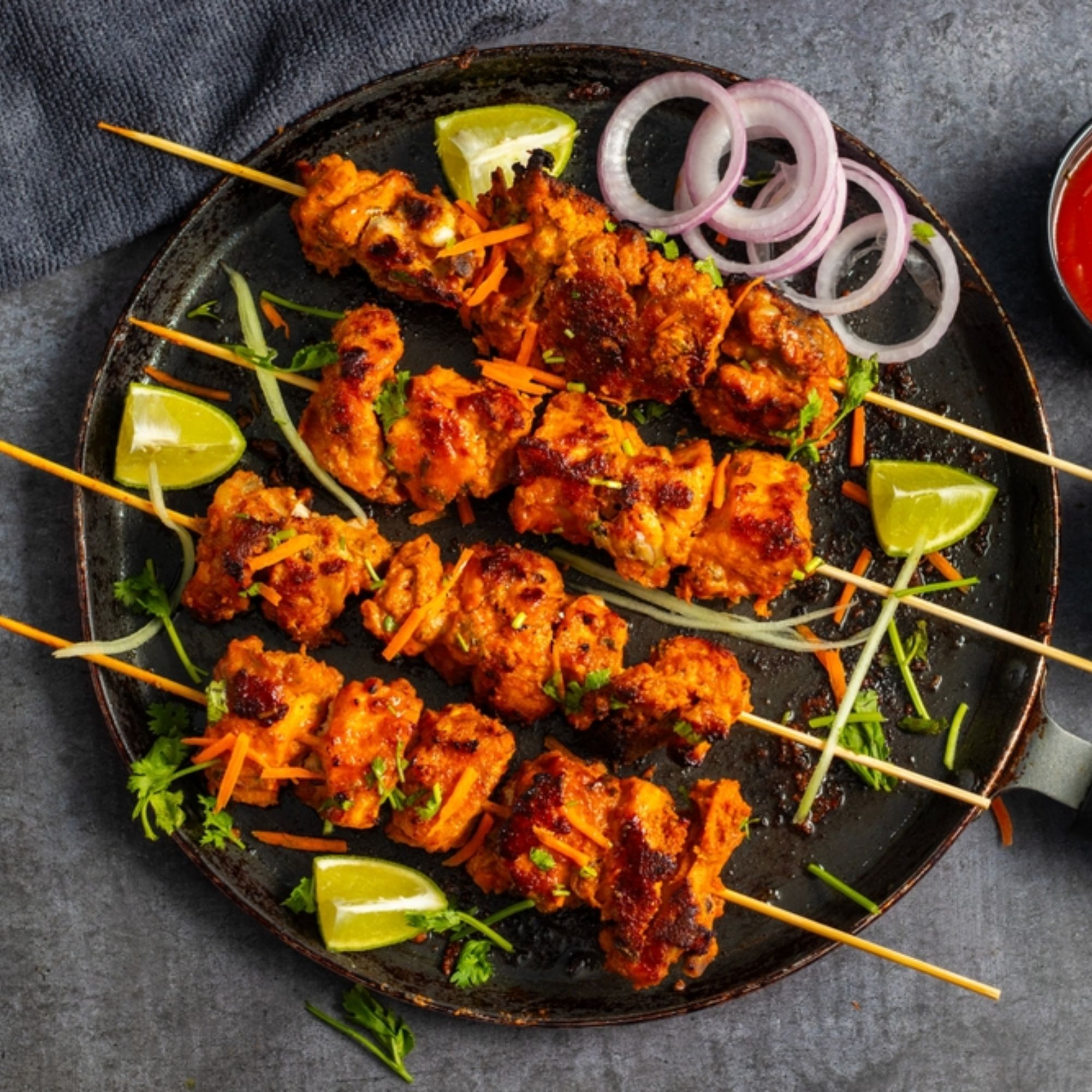 Grilled skewers of marinated chicken on a plate, garnished with sliced onions, carrot strips, lime wedges, and fresh herbs in indian restaurants in franklin tn. A small bowl of red sauce sits nearby.