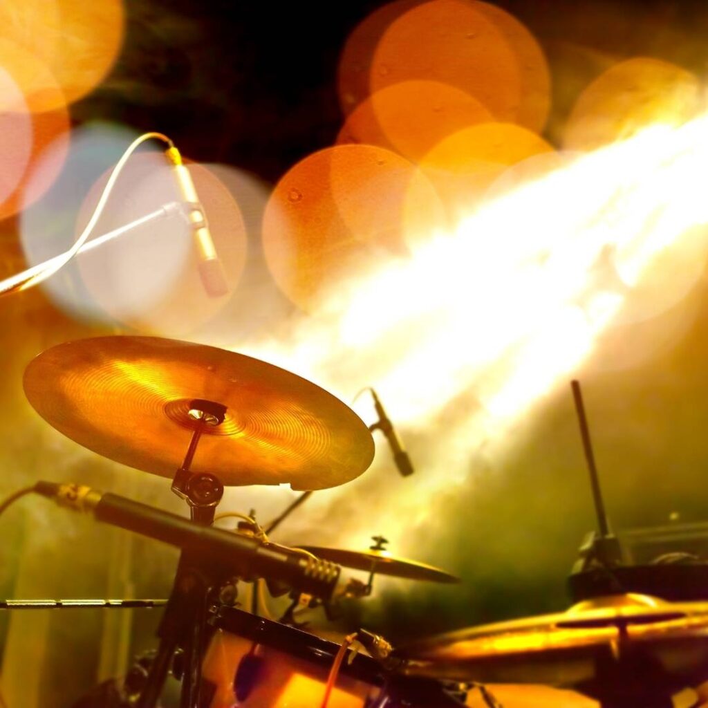 A vibrant stage scene with a glowing cymbal, microphone, and drum set under warm yellow lighting. The atmosphere is energetic, suggesting live music.